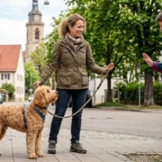 Eine Hundehalterin steht mit ihrem angeleinten, goldfarbenen Doodle auf einem Gehweg in Stuttgart und signalisiert einer passierenden Person mit einer freundlichen, stoppenden Handbewegung, dass der Hund Abstand benötigt. Im Hintergrund ist eine typische Stuttgarter Wohnstraße mit einem Kirchturm zu sehen. Das Bild verdeutlicht das Thema Individualdistanz und dass man seinen Hund nicht streicheln lassen muss.